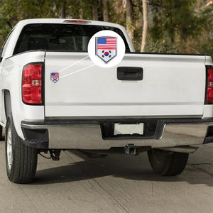 Xalvoria dual flag emblem sticker featuring United States and South Korea flags applied on a white pickup truck tailgate, shield-shaped chrome-style badge shown in an outdoor driving scene