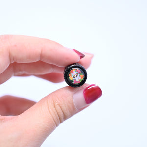 Hand-held view of the sugar skull valve stem cap, showing its compact size and colorful design on a black metal body.