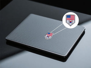 Xalvoria dual flag emblem sticker featuring United States and South Korea flags applied on a laptop surface, shield-shaped chrome-style badge shown in a close-up usage scene
