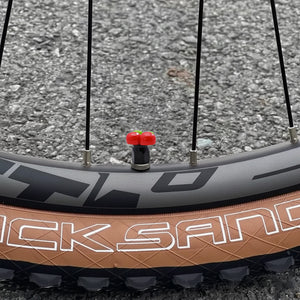 Cherry-shaped valve stem cap installed on a bicycle wheel, showing the real-use look and cute red fruit design on the tire valve.