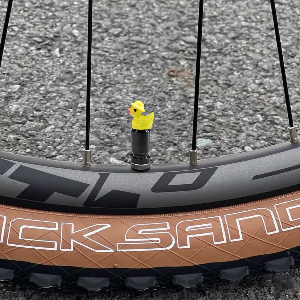 Yellow duck valve stem cap installed on a bicycle wheel, shown outdoors on pavement.