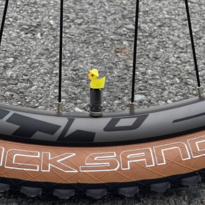 Yellow duck valve stem cap installed on a bicycle wheel, shown outdoors on pavement.